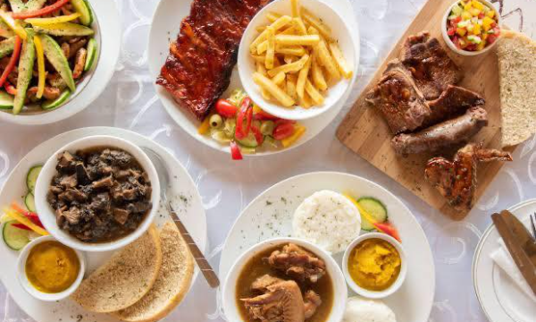 A variety of different dishes. A plate of bbq ribs and french fries with salad, a bowl of green salad, a bowl of stew with two slices of bread, a chicken stew with two portions of pap, lastly a board of sausage, chicken and steak with bread and salad all by Makhelwane -Vilakazi Street Restaurants